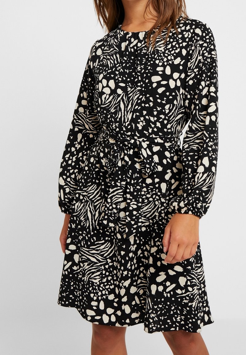 Černé a krémové šaty se vzorem, dlouhými rukávy, páskem v pase a vrstevnatým volánkovým lemem. Obsahují abstraktní tvary a pohodlný siluetu.