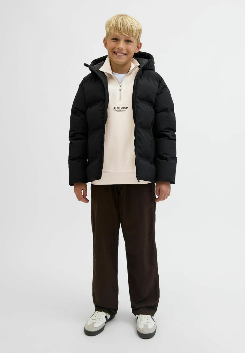 Svart pufferjacka med huva, bärs över en ljus beige zip-up sweatshirt. Bruna löst sittande byxor och vita skor med grå detaljer.