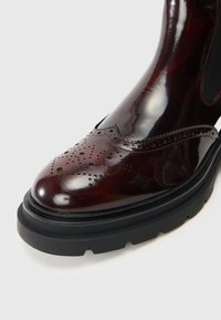 Stivale in pelle mogano lucida con dettagli brogue, punta tonda, suola nera a contrasto e cuciture lungo il bordo superiore.