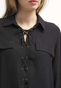 Blouse noire avec col, devant lacé avec œillets et deux poches poitrine. Fabriquée en tissu doux et lisse, avec une coupe décontractée.