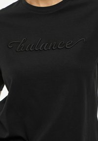 Svart bomulls T-shirt med en texturerad svart broderad "balance"-logotyp över bröstet, rund halsringning och korta ärmar.