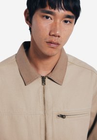 Giacca beige con zip e colletto beige, dotata di una cerniera frontale e di una piccola tasca sul petto con cerniera. Tessuto con texture liscia.
