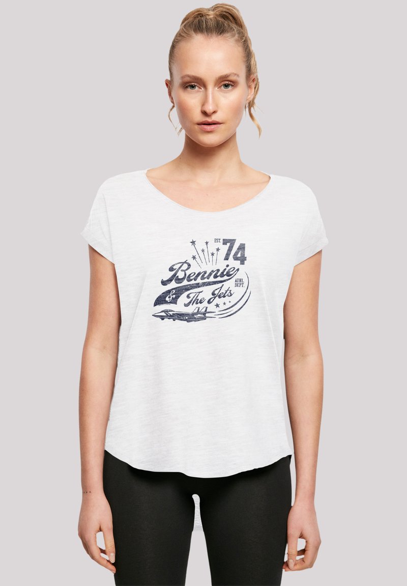 Eine Frau steht selbstbewusst da, trägt ein weißes T-Shirt mit einem Grafikdesign, das "Bennie & The Jets" und ein Flugzeugbild zeigt, und kombiniert es mit schwarzen Leggings.