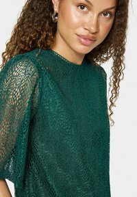 Haut en dentelle verte, avec des motifs ouverts complexes, des manches courtes bouffantes et un col haut. Texture subtile et apparence légère.
