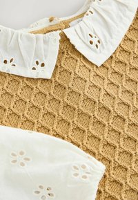 Haut en tricot présentant un motif texturé couleur miel avec un col brodé blanc et des découpes florales le long des bords.