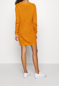 Vestito in maglia a coste di colore arancione brillante, con scollo rotondo, maniche lunghe e texture lavorata a maglia, abbinato a sneakers bianche.