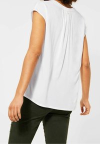 Persona che indossa una semplice blusa bianca a maniche corte e pantaloni verdi scuri, vista da dietro su uno sfondo bianco semplice.