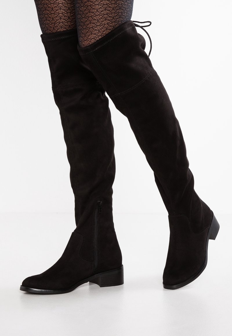 Bottes noires en daim au-dessus du genou avec talons bas, fermeture éclair latérale et lacet à l'arrière, portées avec des collants à motifs.