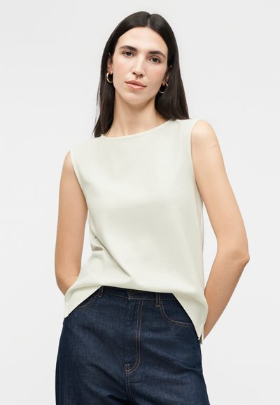 Γυναίκα με μακριά σκούρα μαλλιά, φορώντας χρυσούς κρίκους στα αυτιά, λευκό αμάνικο τοπ και σκούρο μπλε ψηλόμεσα τζιν, σε ουδέτερο φόντο.