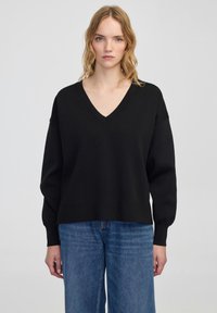 Zwarte V-hals trui met lange mouwen, gemaakt van een zachte stof. Ontspannen pasvorm en geribbelde zoom. Gemaakt met lichte blauwe wijde jeans.