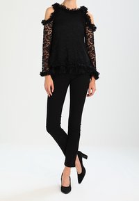 Svart spetsblus med kallaxel-design, volangkrage och manschetter; kombinerad med svarta tajta byxor och högklackade skor.