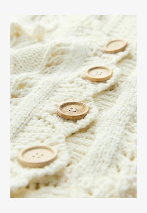 Pletena krem svijetla pulover s teksturiranim uzorcima, sa pet drvenih gumba s četiri rupe svaki duž prednje strane.