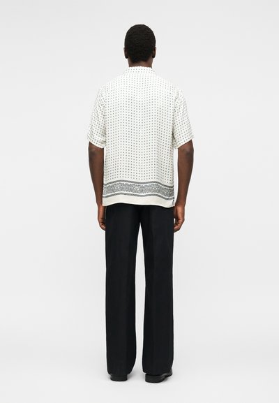 Άνδρας που φοράει λευκό πουκάμισο με κοντά μανίκια με μικρά μαύρα σχέδια και διακοσμητική μαύρη μπορντούρα, σε συνδυασμό με φαρδιά μαύρα παντελόνια, όρθιος.