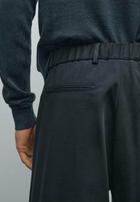 Close-up of a person wearing dark, elastic-waist trousers with a back welt pocket, paired with a dark long-sleeve top.