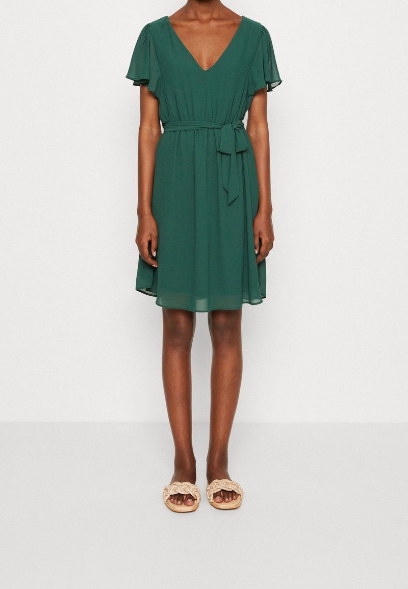 Robe verte à col en V avec manches courtes évasées, taille ceinturée et jupe fluide. Associée à des sandales beige à enfiler avec un design tressé.