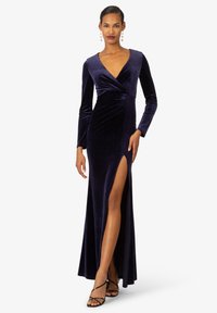 Navy Blau Samt-Abendkleid mit langen Ärmeln, tiefem Ausschnitt, Raffung und einem slits bis zum Oberschenkel; kombiniert mit schwarzen Sandalen mit Riemchen.