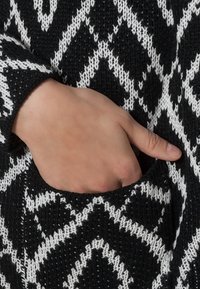 Jersey de punto en blanco y negro con un patrón en zigzag; cuenta con un bolsillo frontal y puños acanalados, mostrando un tejido texturizado.