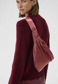 Rózsasz�ín nylon sling táska puha, gyűrött textúrával és széles selyempánttal, egy bordó pulóver fölött viselve, egyszínű háttér előtt.