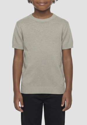 Dijete odjeveno u svijetlosivi kratki pleteni džemper i crne hlače, stoji nasuprot jednobojnoj svijetloj pozadini.