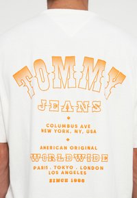 White short-sleeve t-shirt with orange graphic text reading "TOMMY JEANS" and location details. Text features a bold, textured design.