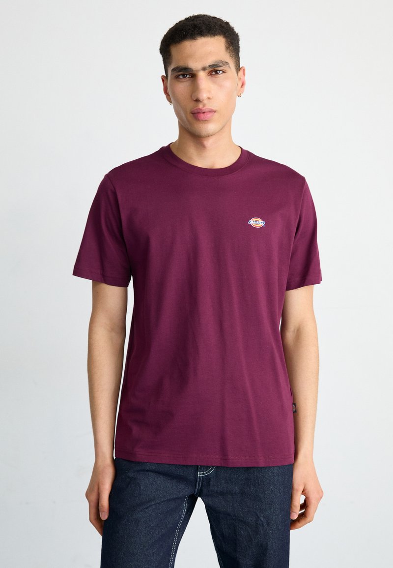 T-shirt in cotone bordeaux con maniche corte, scollatura rotonda e piccolo logo ricamato sul lato sinistro del petto, abbinato a jeans scuri.