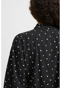 Blusa nera con un piccolo motivo a cuori color crema. Presenta dettagli arricciati alle spalle e un collo a volant. Tessuto dal tocco liscio.