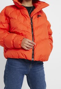 Orange puffer jacket with a high collar, featuring a black zipper and Nike logo. Ribbed cuffs and a padded design with a glossy finish.