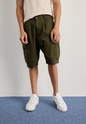 Pantalones cortos cargo de algodón verde oliva, con un corte relajado, dos bolsillos laterales y dobladillos enrollados, combinados con zapatillas blancas.