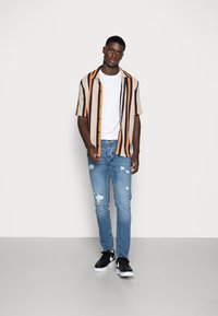 Kurzärmliges, gestreiftes Button-up-Hemd in Beige, Marineblau und Orange; weißes T-Shirt darunter; hellblaue, ausgewaschene Jeans; schwarze Sneakers.