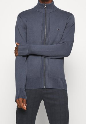 Homme portant un pull zippé en tricot bleu à col montant et un pantalon à carreaux foncé, debout les bras croisés.