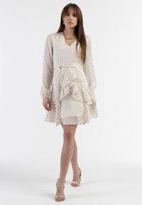 Rochie de culoare crem deschis, cu mâneci lungi, tiv volanat și un model texturat subtil. Complettată de sandale cu barete bej.