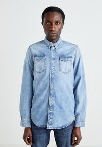Camisa de mezclilla azul claro con cuello, mangas largas, cierre de botones en la parte delantera y dos bolsillos en el pecho. Textura suave con un diseño casual.