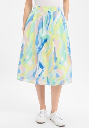 Femme portant une jupe mi-longue avec des motifs abstraits aux coups de pinceau bleu, vert, jaune et rose pastel, baskets blanches et haut blanc.