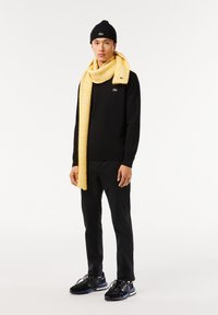 Maglione nero, sciarpa a coste gialla, berretto nero, pantaloni neri e sneakers nere con suola texturizzata. Logo Lacoste su maglione e sciarpa.