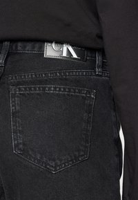 Juodi džinsai su išskirtine stačiakampio formos užpakaline kišene ir tekstūruotu juosmens etiketės ženkleliu, ant kurio yra "Calvin Klein Jeans" logotipas.