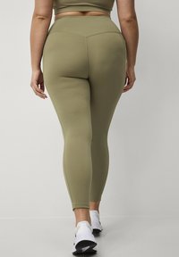 Olijfgroene high-waisted leggings van rekbare stof, met een gladde textuur en een aansluitende snit, gecombineerd met witte sneakers.