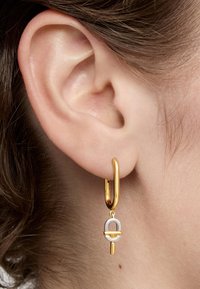 Złoty kolczyk w kształcie obręczy z zawieszką w srebrze i złocie, charakteryzującą się okrągłym wzorem i pionowym słupkiem. Gładka tekstura; nowoczesny geometryczny kształt.