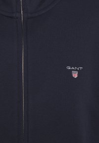 Felpa con zip color navy realizzata in tessuto morbido, dotata di zip argentata e logo GANT ricamato con scudo. Design semplice, senza motivi o trame.