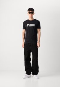 Black t-shirt with a logo print, paired with black loose-fit pants and white sneakers. Model wears dark sunglasses. Minimalist design.