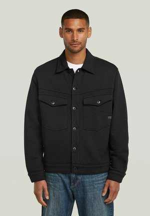 Mann trägt eine schwarze Jacke mit Knöpfen und Klappentaschen, steht vor einem schlichten hellen Hintergrund und hat einen neutralen Gesichtsausdruck.