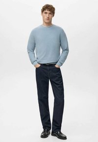 Jasnoniebieski sweter z dzianiny z okrągłym dekoltem, ciemne dżinsy i czarne skórzane buty. Model stoi wyprostowany, z rękami w kieszeniach, na neutralnym tle.