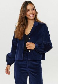 Navy modra žametna jakna s ovratnikom, dvema gumboma in napihnjenimi rokavi. Ujemajoče hlače dopolnjujejo celotno oblačilo.
