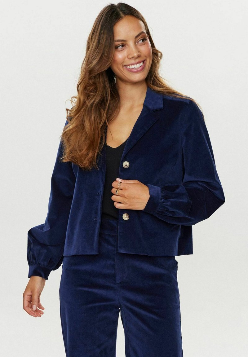 Navy modra žametna jakna s ovratnikom, dvema gumboma in napihnjenimi rokavi. Ujemajoče hlače dopolnjujejo celotno oblačilo.