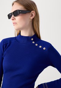 Blå ribbad stickad topp med hög krage, silverknappar längs axeln och subtila texturerade detaljer vid ärmsluten.