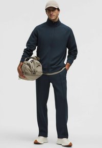 Marineblå zip-up jakke og matchende bukser, lavet af blødt stof. Lysfarvet cap og beige taske med snorelukninger. Hvide sneakers.