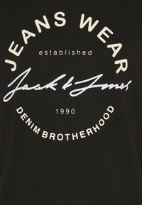 Camiseta negra con un logotipo circular blanco que incluye el texto "JEANS WEAR", "Jack & Jones" y "DENIM BROTHERHOOD" con el año de establecimiento 1990.