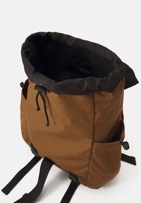 Mochila de tela marrón con cierre en la parte superior de color negro y rollo, cordón ajustable, bolsillo lateral y correas dobles negras en la parte inferior.