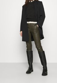 Černý dlouhý kabát přes černou mikinu, olivově zelené kožené kalhoty a černé kozačky ke kolenům s texturovaným povrchem.