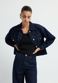 Žena v tmavě modré džínové bundě a kalhotách se zlatými knoflíky, černém topu a zlatých naušnicích, s rukama na bocích, looking dolů.