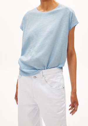 Hellblaues Kurzarm-Top, in weiße Jeans mit hoher Taille gesteckt, an einer Person mit neutralem Hintergrund.